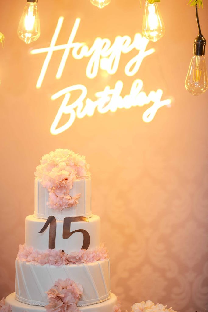 A beautiful three-tiered cake adorned with flowers against a neon 'Happy Birthday' sign.